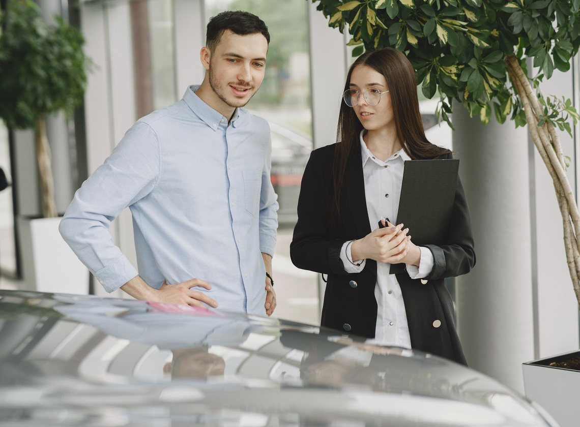 Consultora de blazer preto conversando com um cliente de camisa azul-clara diante do capô de um carro